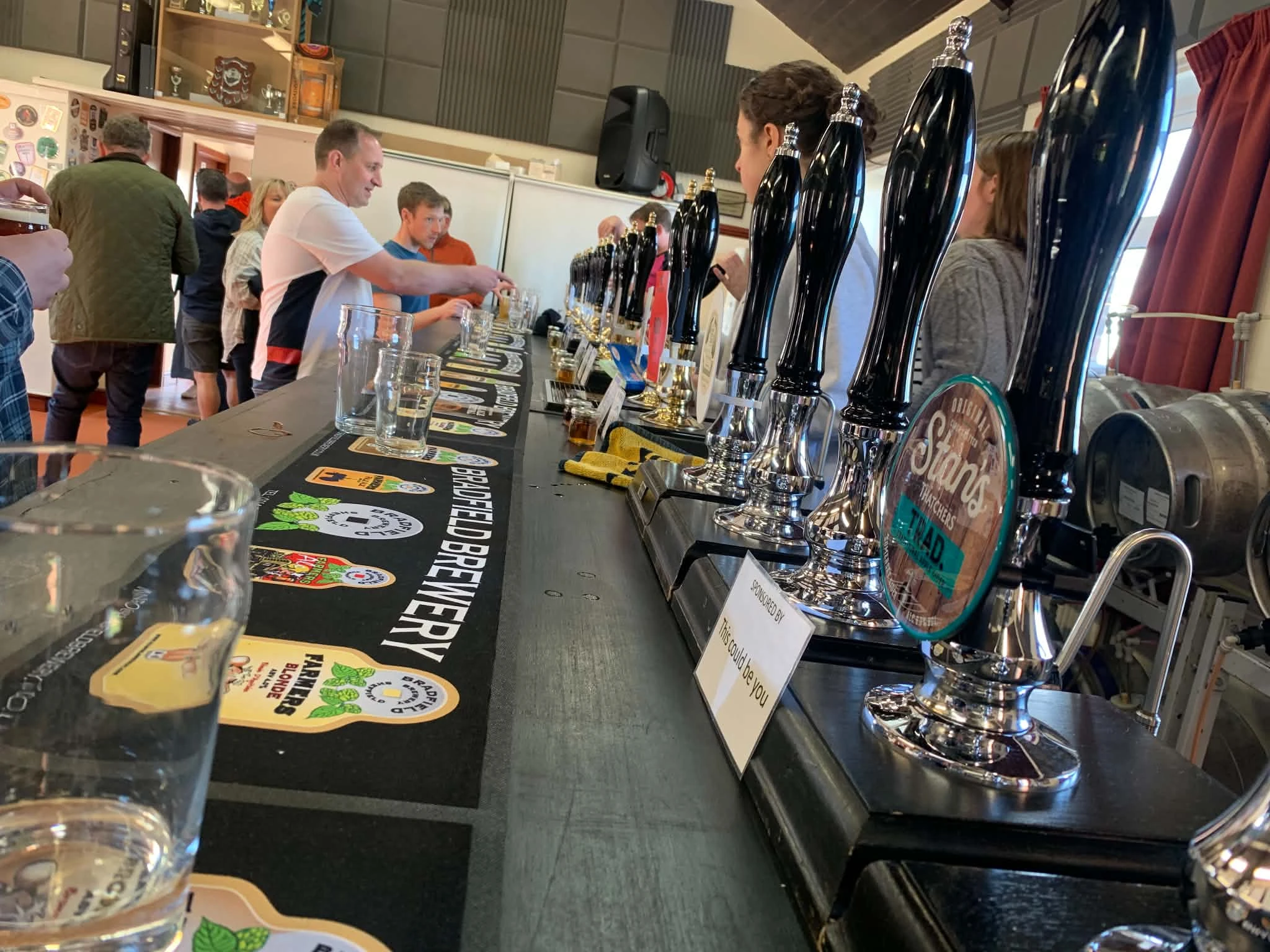 A row of hand-pulled beer pumps with people at the bar during the Emley Real Ale Festival.