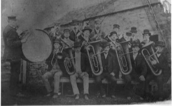 An early black-and-white photograph of band members with brass instruments and a bass drum outside a stone building.