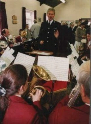A conductor standing in front of seated band members during a rehearsal, with percussion instruments behind him.