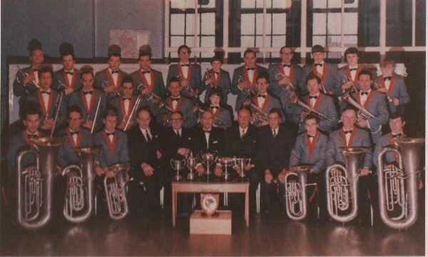 Emley band members in red uniforms with brass instruments and trophies arranged on a table.